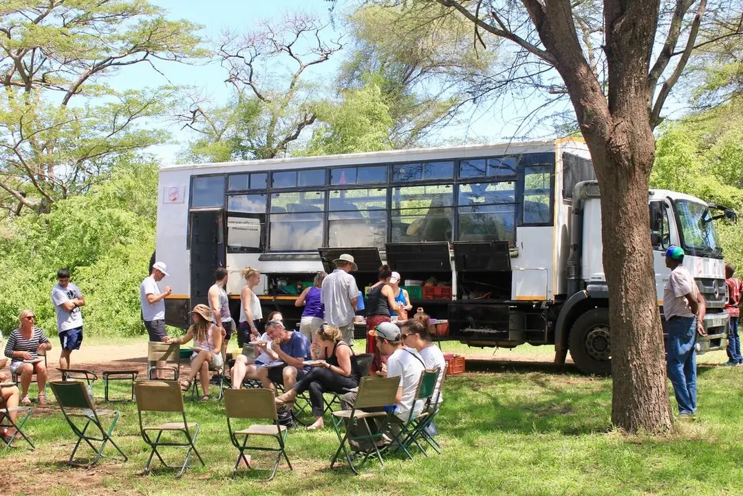 Tour group eats lunch on camping chairs next to overland vehicle under tree.