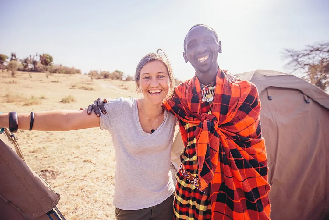 Female traveller and male Masai warrior smile as they pose in front of canvas tent.