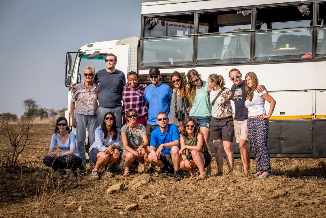 Group of travellers pose in front of overland vehicle on rocky ground.