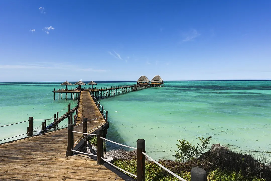 Wooden walkway built above tropical sea leading to thatch huts.