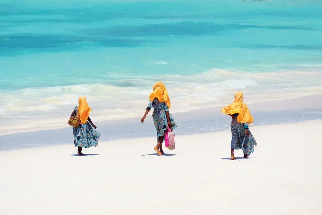 Three local women walk along bright white sandy beach with turquoise blue ocean.